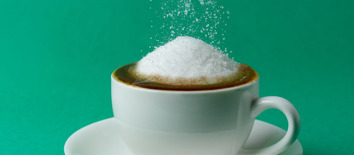 Cup full of sugar, too much, overeating. Hand with spoon adding more sugar to coffee